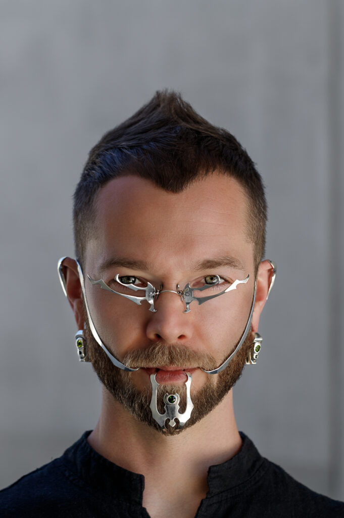 Jeramie Bellmay of Atavus Couture looking into the camera wearing custom sterling silver jaw jewelry, spectacles, ear cuffs, and jeweled weights